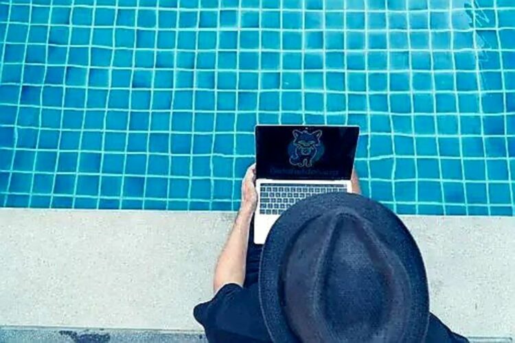 Una persona con sombrero negro sostiene un portátil al borde de una piscina