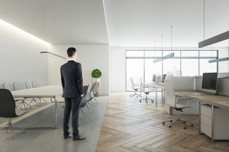 Pensive young businessman in suit standing in wooden coworking meeting room interior with daylight, furniture and equipment. Design and ceo worker concept